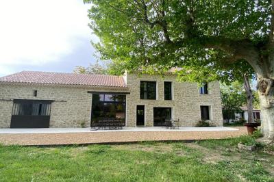 Maison avec terrasse entourée de végétation, située dans un jardin avec allée en gravier et ciel dégagé en arrière-plan.