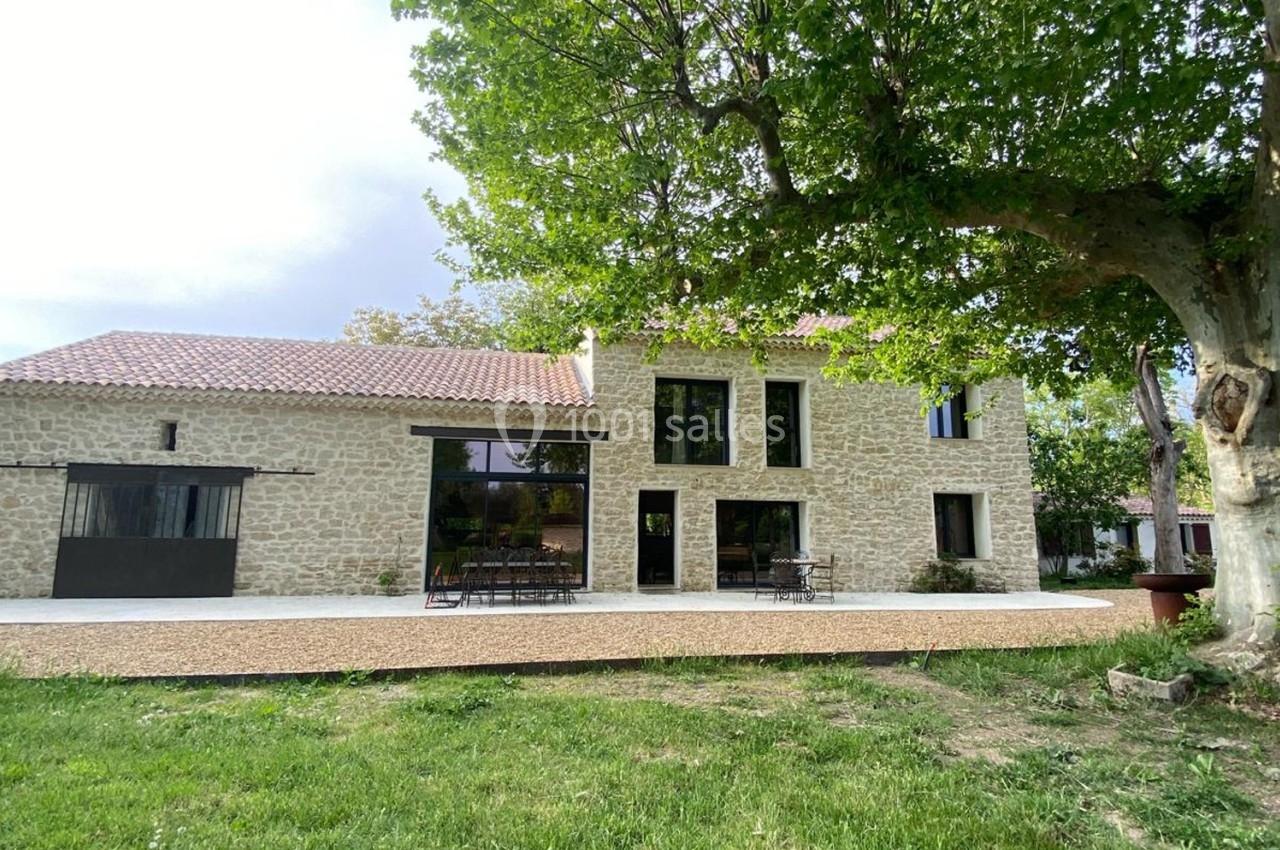 Façade d'une maison en pierre avec grandes baies vitrées, terrasse aménagée et arbre au premier plan.