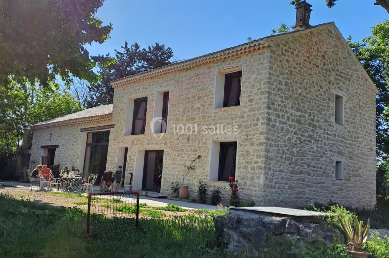 Maison en pierre avec grandes fenêtres, terrasse aménagée et personnes assises autour d'une table dans un jardin verdoyant.