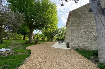 Maison avec terrasse entourée de végétation, située dans un jardin avec allée en gravier et ciel dégagé en arrière-plan.