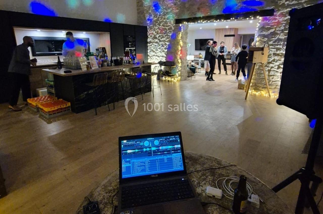 Vue d'une salle de réception avec un DJ en arrière-plan, des invités discutant et un bar illuminé.