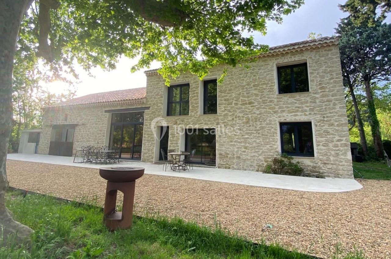 Façade d'une maison en pierre avec terrasse, mobilier de jardin et allée gravillonnée, entourée de verdure.