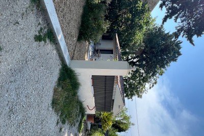 Maison avec terrasse entourée de végétation, située dans un jardin avec allée en gravier et ciel dégagé en arrière-plan.