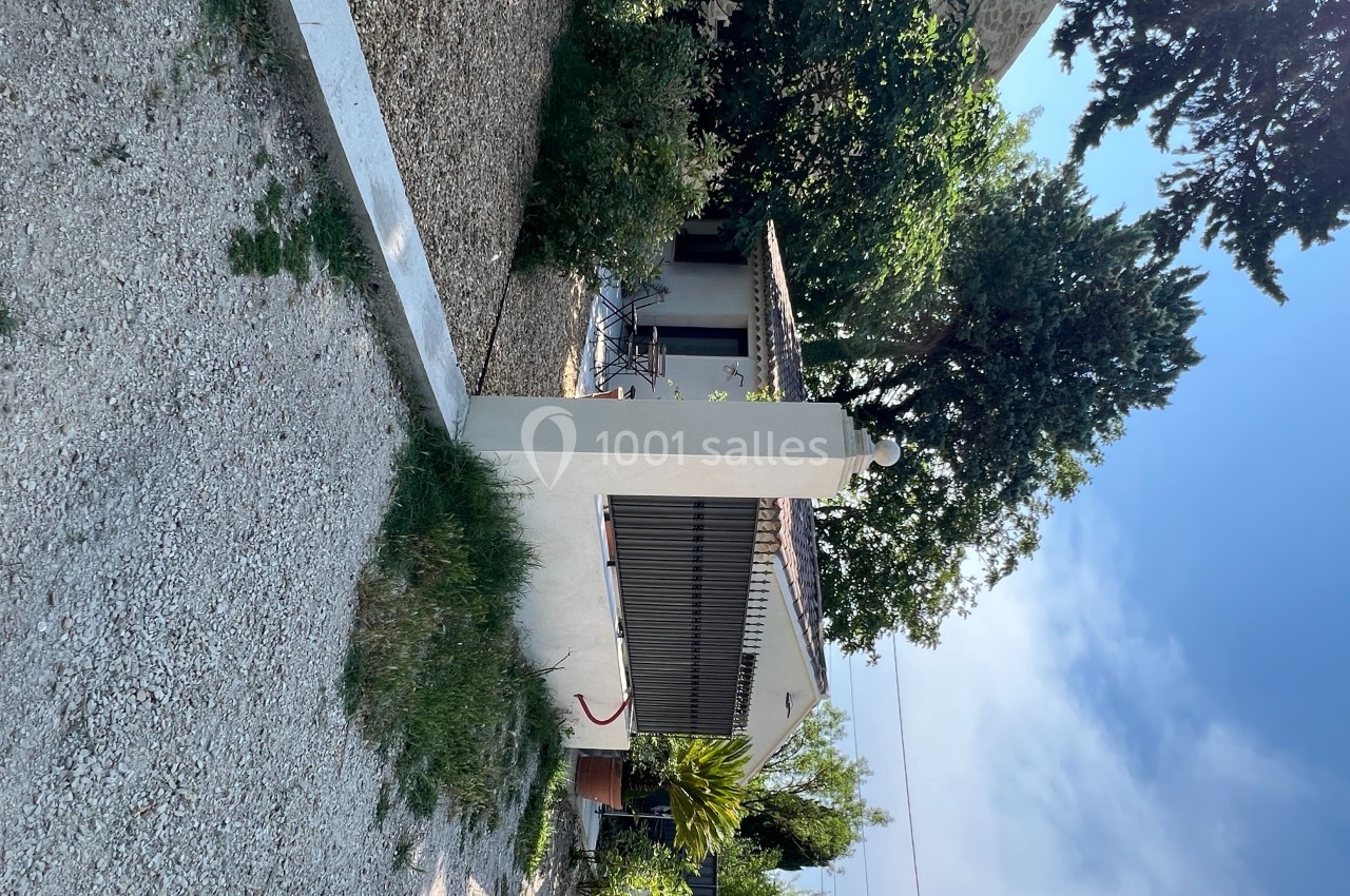 Maison avec terrasse entourée de végétation, située dans un jardin avec allée en gravier et ciel dégagé en arrière-plan.