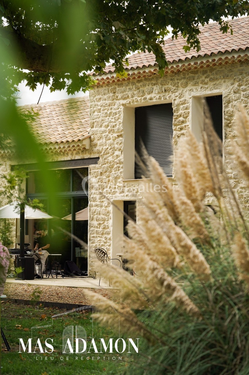 Façade en pierre d'un bâtiment avec grandes fenêtres, terrasse aménagée et végétation environnante.
