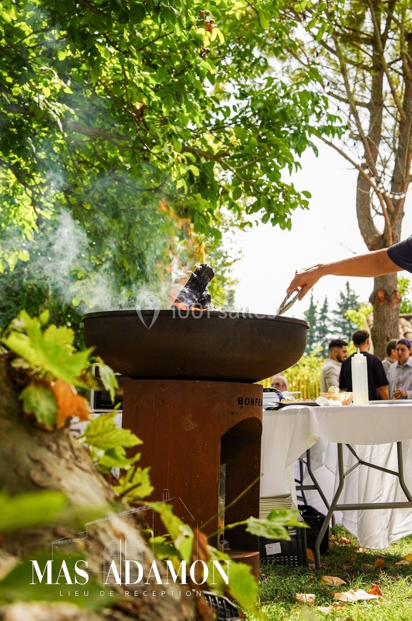Personne cuisant des aliments sur un brasero en plein air, entourée de verdure et de convives en arrière-plan.
