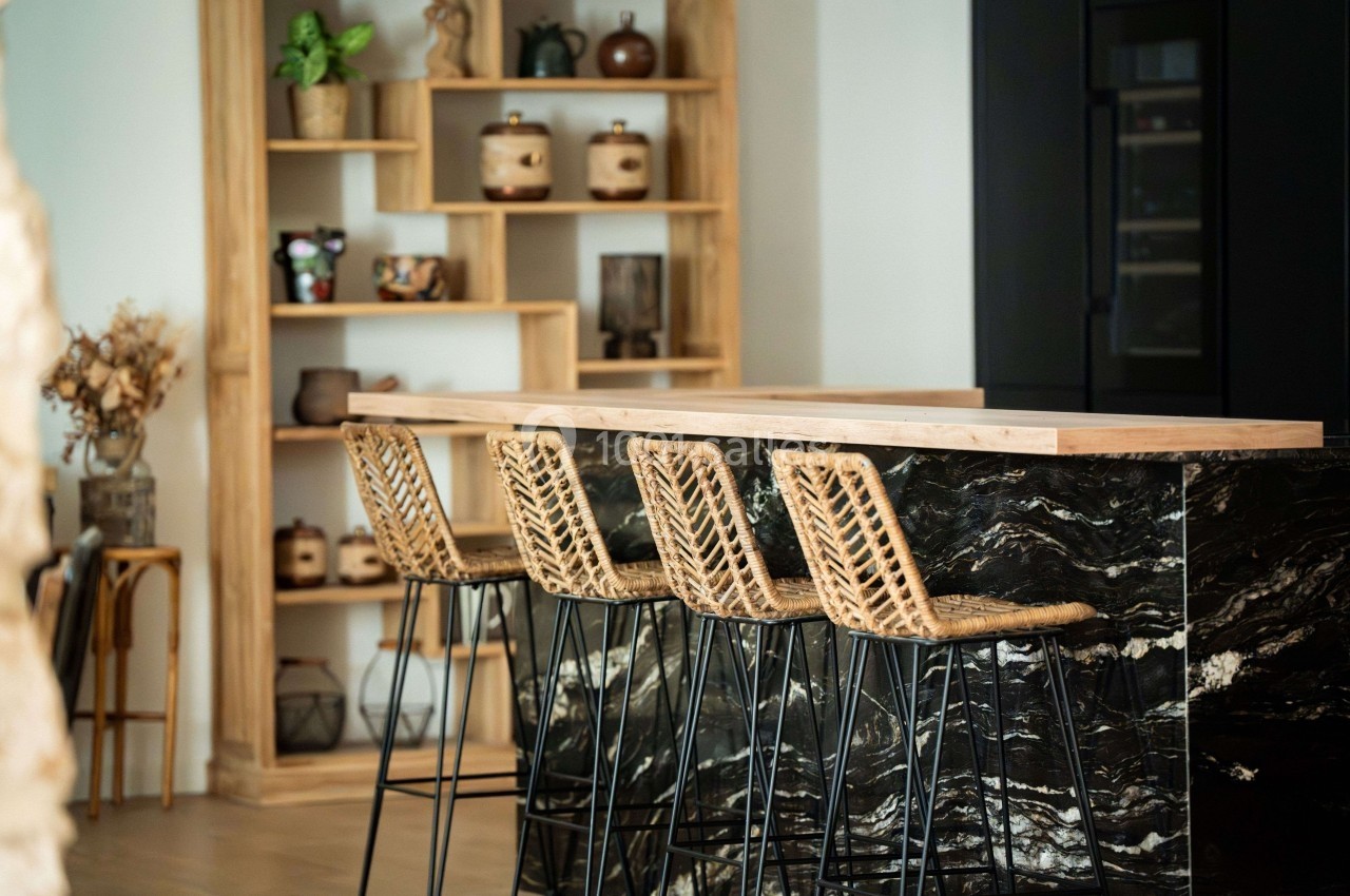 Quatre tabourets en rotin alignés devant un comptoir en marbre noir dans une cuisine moderne avec étagères en bois.