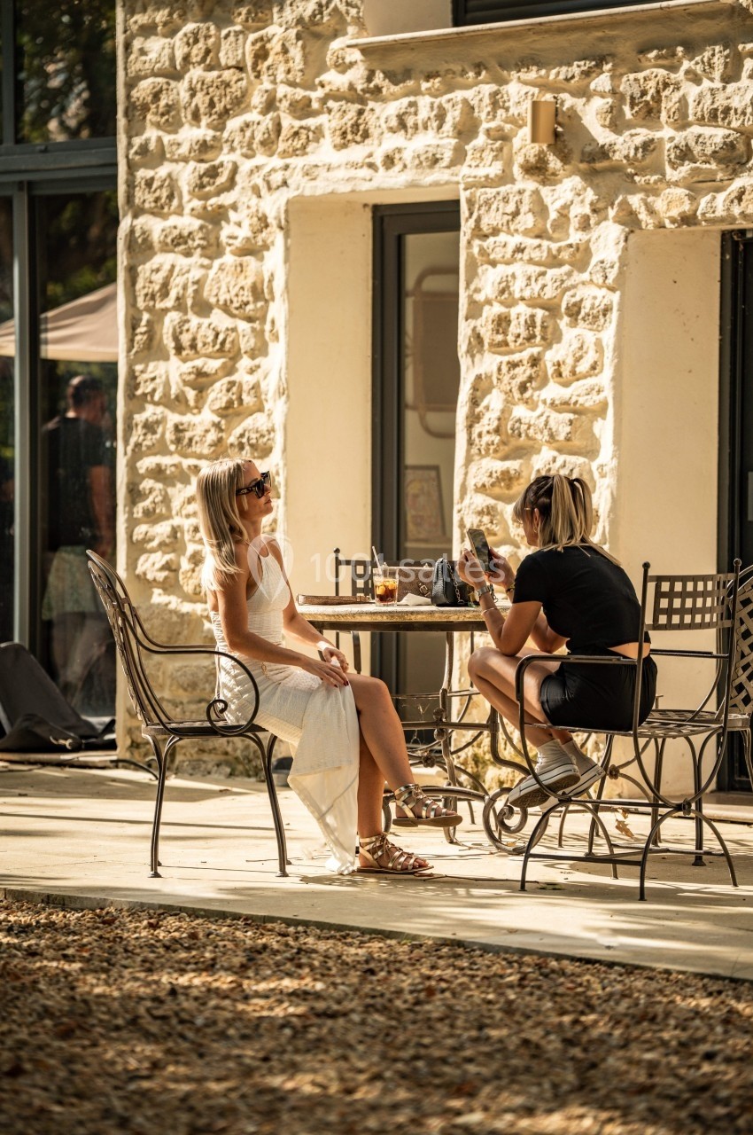 Deux femmes discutent à une table en terrasse, devant un bâtiment en pierre baigné de lumière naturelle.