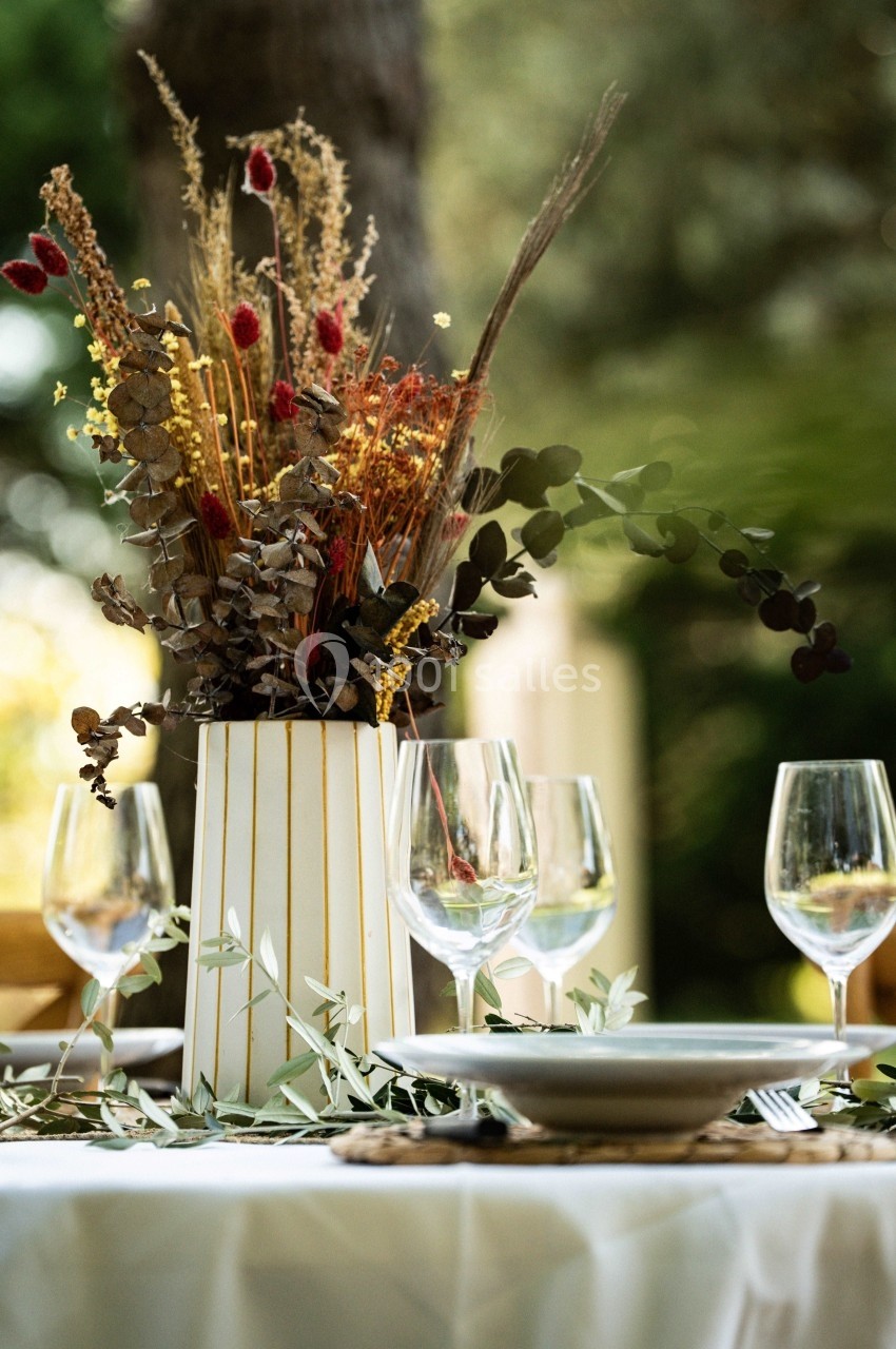 Centre de table avec bouquet de fleurs séchées dans un vase rayé, entouré de verres et assiettes sur une nappe blanche.
