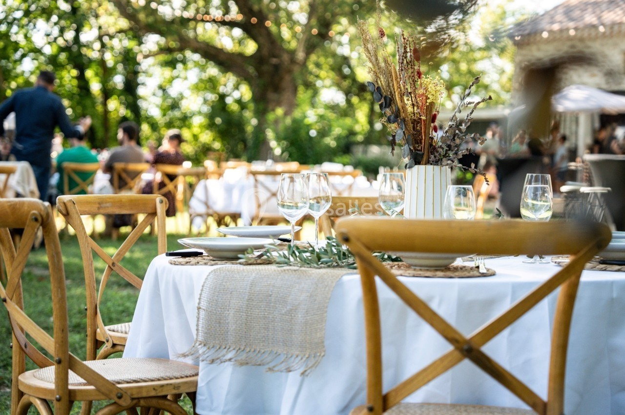 Table dressée en extérieur avec vaisselle, verres et décoration florale, entourée de chaises en bois dans un jardin.