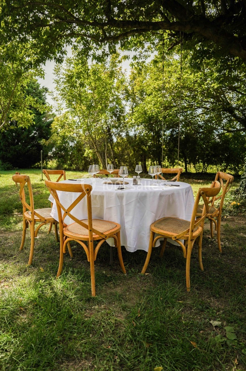 Table ronde dressée avec une nappe blanche et des verres, entourée de chaises en bois, sous un arbre dans un jardin…