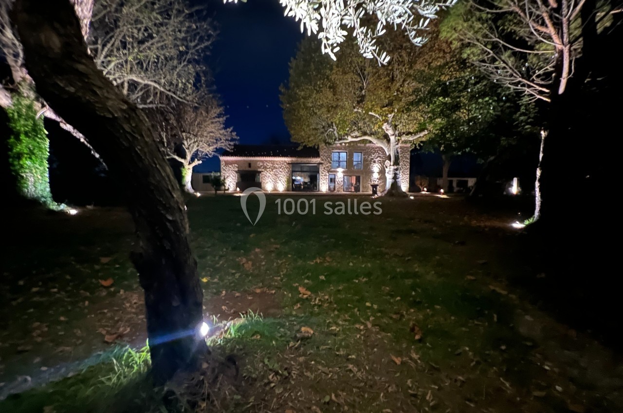 Maison en pierre éclairée la nuit, entourée d'arbres et d'un jardin sombre avec des lumières au sol.