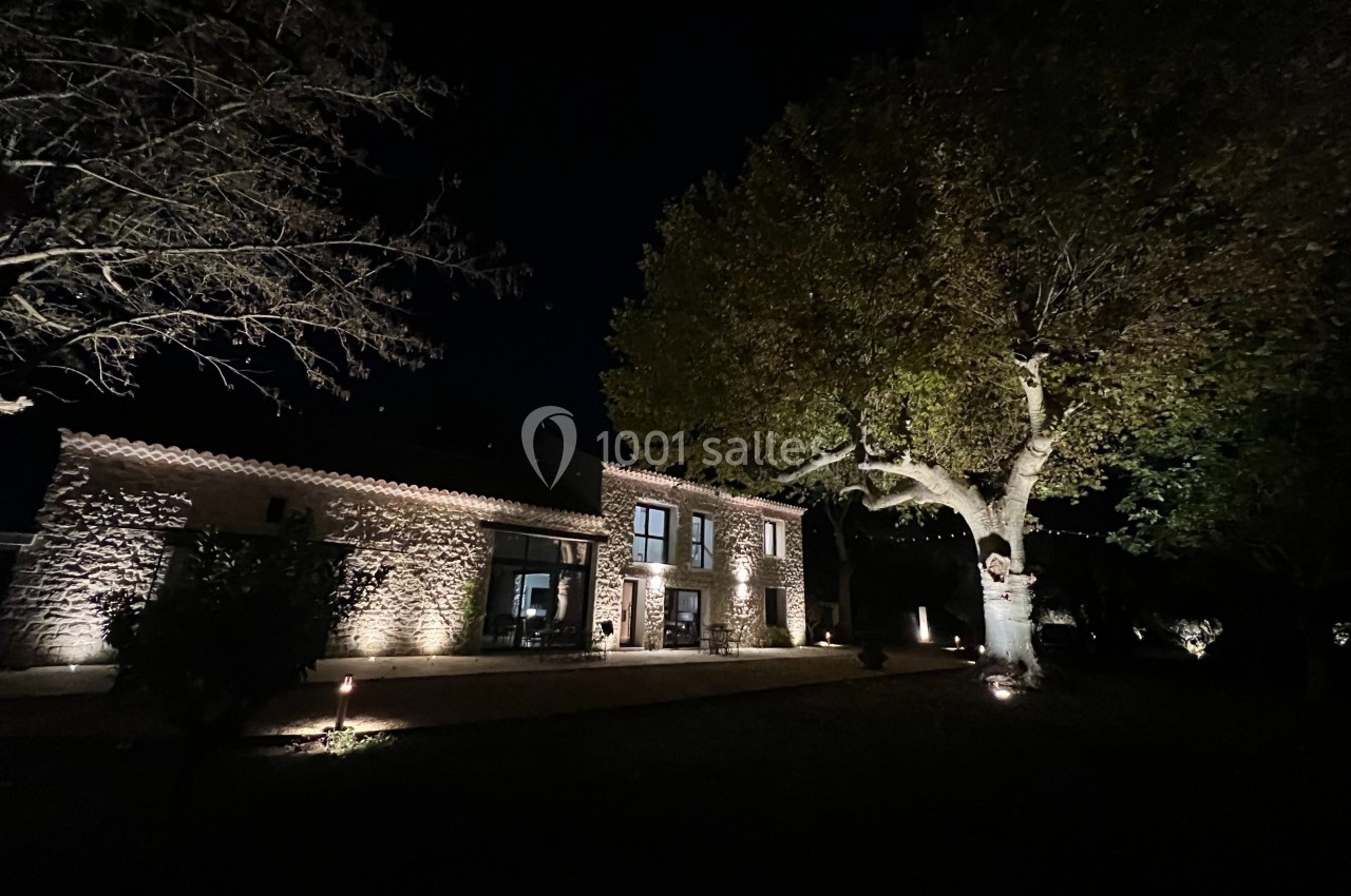 Façade d'une maison en pierre éclairée la nuit, entourée d'arbres et d'un jardin sombre.