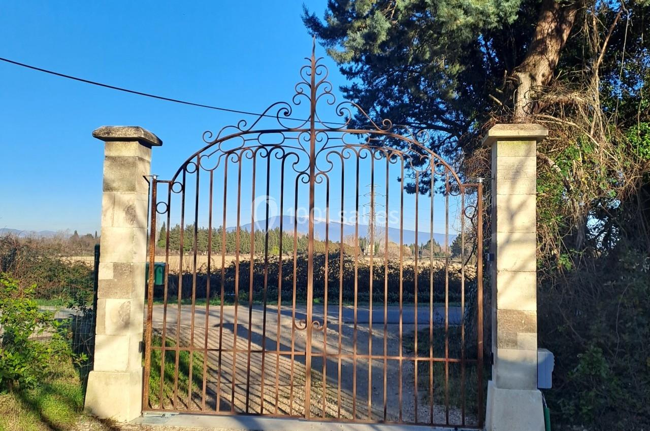 Portail en fer forgé orné de motifs, encadré par deux piliers en pierre, donnant sur un chemin rural.