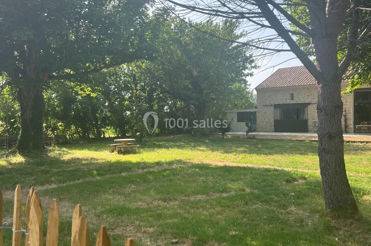 Jardin verdoyant avec pelouse, arbres et une maison en pierre au fond, vu à travers une clôture en bois.