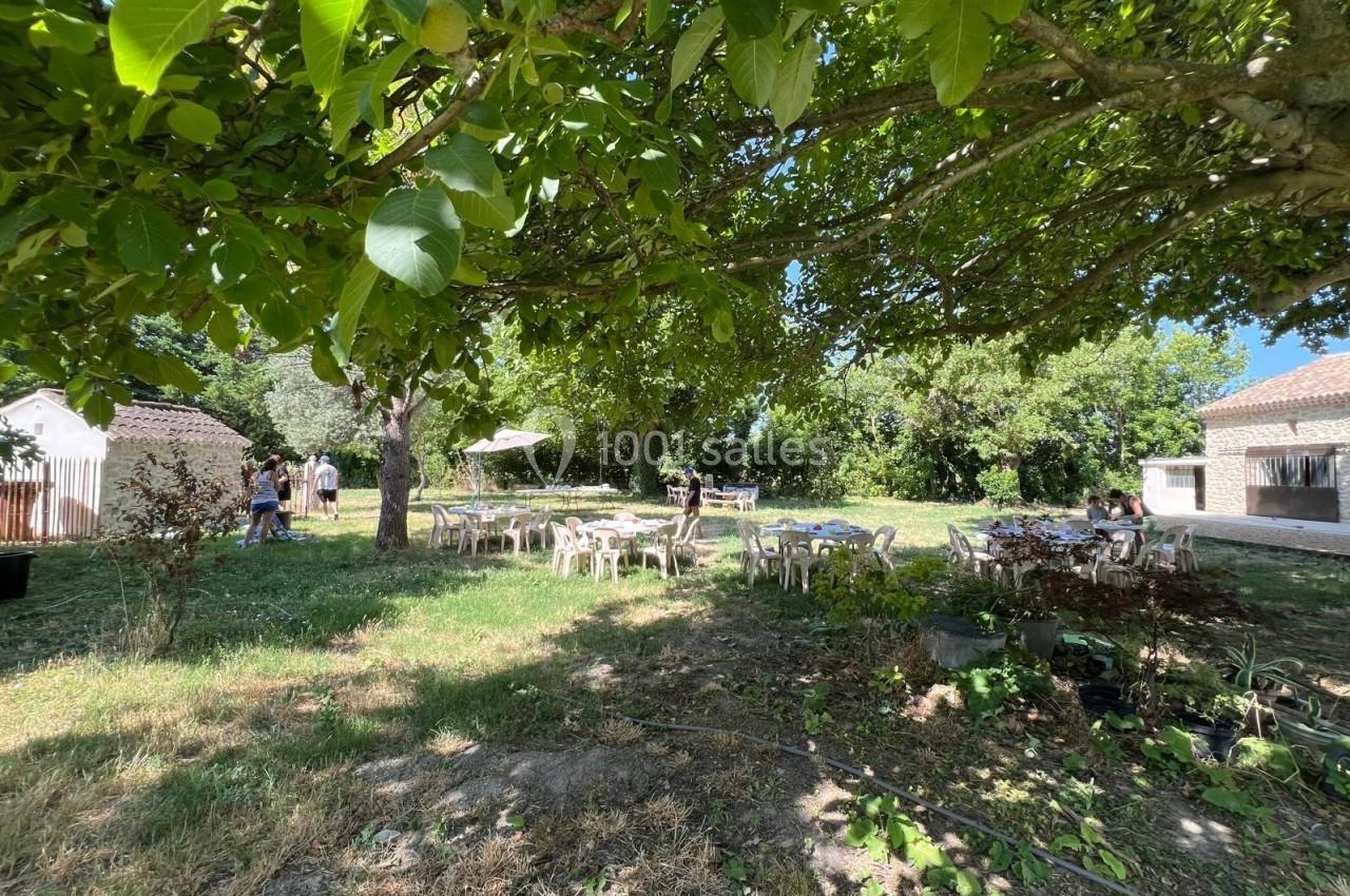 Des tables et chaises blanches disposées sous des arbres dans un jardin ombragé, avec des bâtiments en arrière-plan.