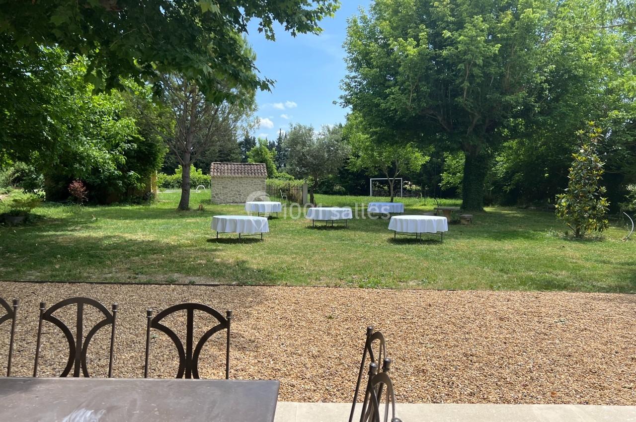 Jardin avec des tables rondes dressées sous des arbres, entouré de verdure et d'un sol en gravier au premier plan.