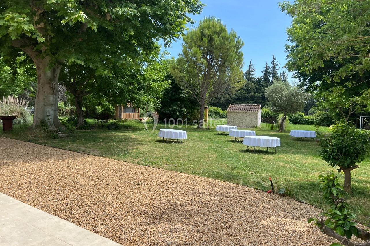 Jardin verdoyant avec des tables rondes couvertes de nappes blanches, entouré d'arbres et d'une allée en gravier.