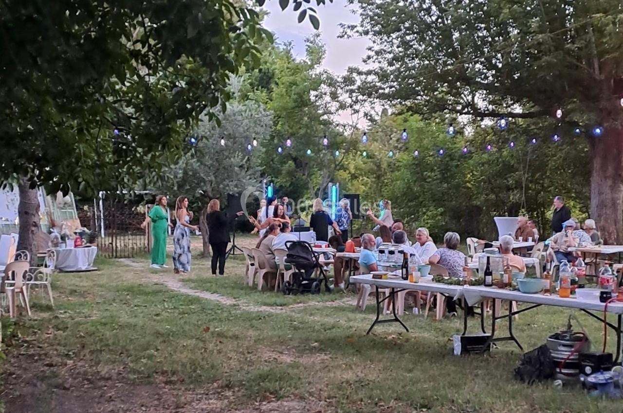 Personnes réunies en extérieur pour un repas festif, avec tables dressées, guirlandes lumineuses et arbres en arrière-plan.