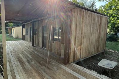 Maison avec terrasse entourée de végétation, située dans un jardin avec allée en gravier et ciel dégagé en arrière-plan.