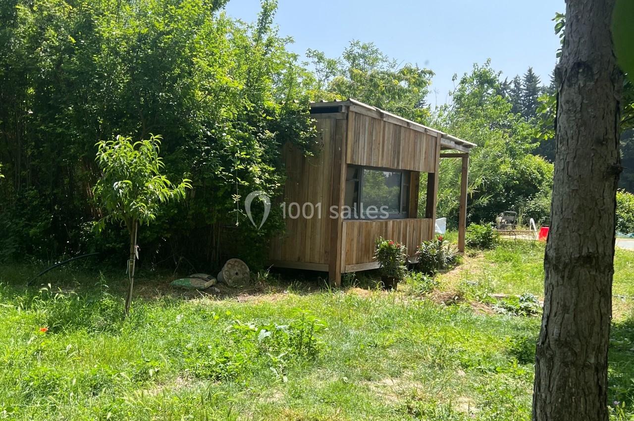 Cabane en bois entourée de végétation dense dans un jardin ensoleillé avec des arbres et de l'herbe.