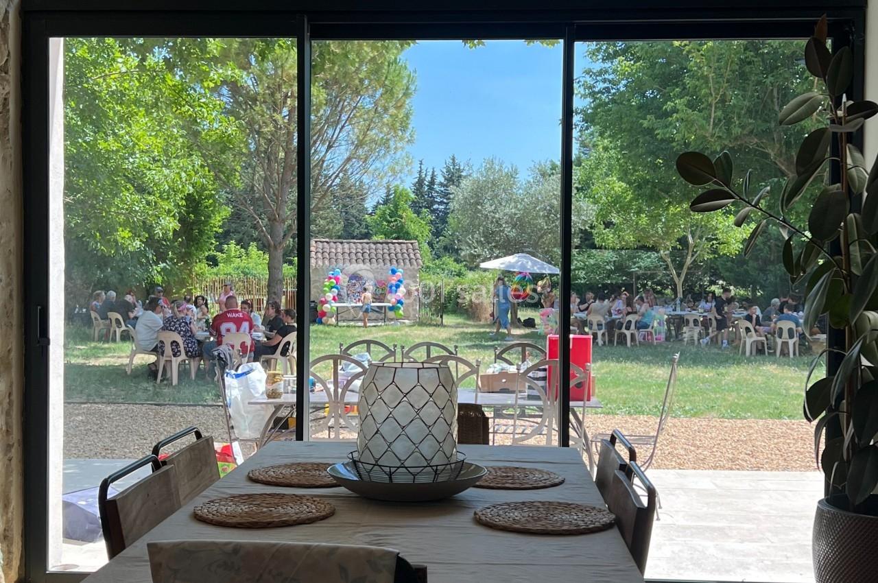 Vue depuis une salle à manger sur un jardin ensoleillé avec des tables et des personnes réunies à l'extérieur.