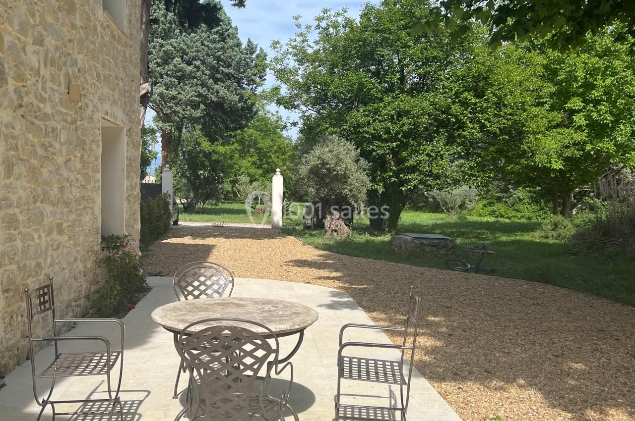Terrasse en pierre avec table et chaises en métal, entourée de verdure et bordée d'un chemin gravillonné.