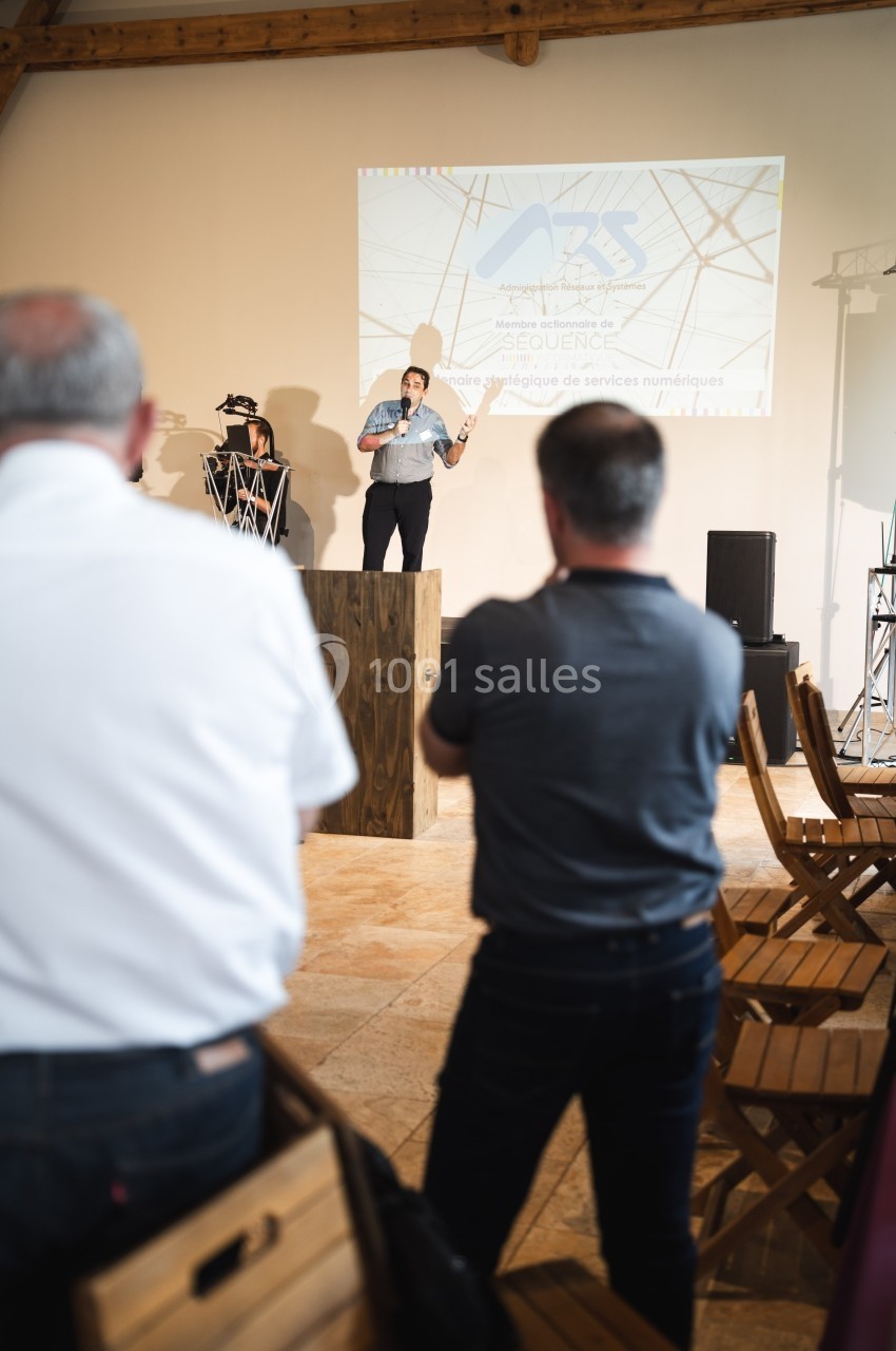 Une personne présente sur scène devant un public dans une salle avec projection d'une diapositive en arrière-plan.