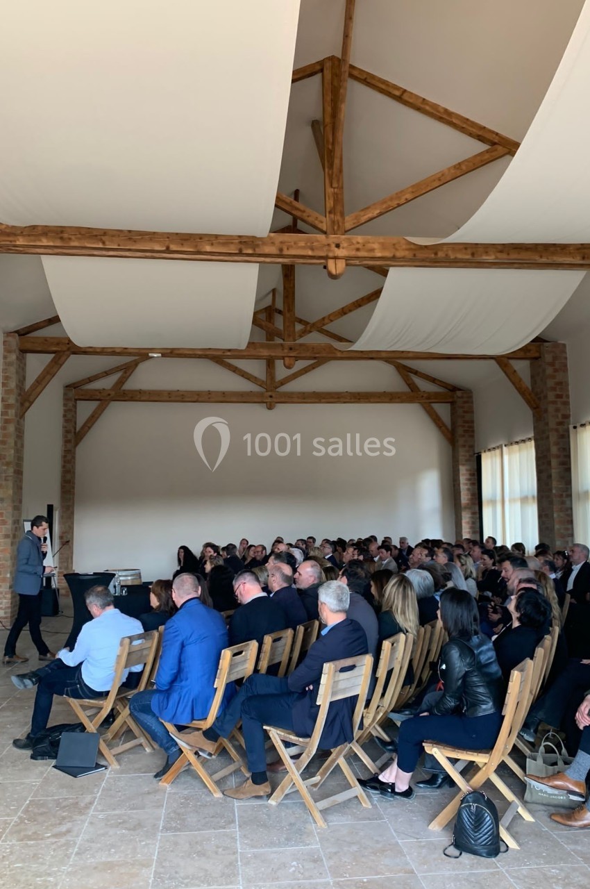 Un conférencier s'adresse à un public assis dans une salle lumineuse avec une charpente en bois apparente.