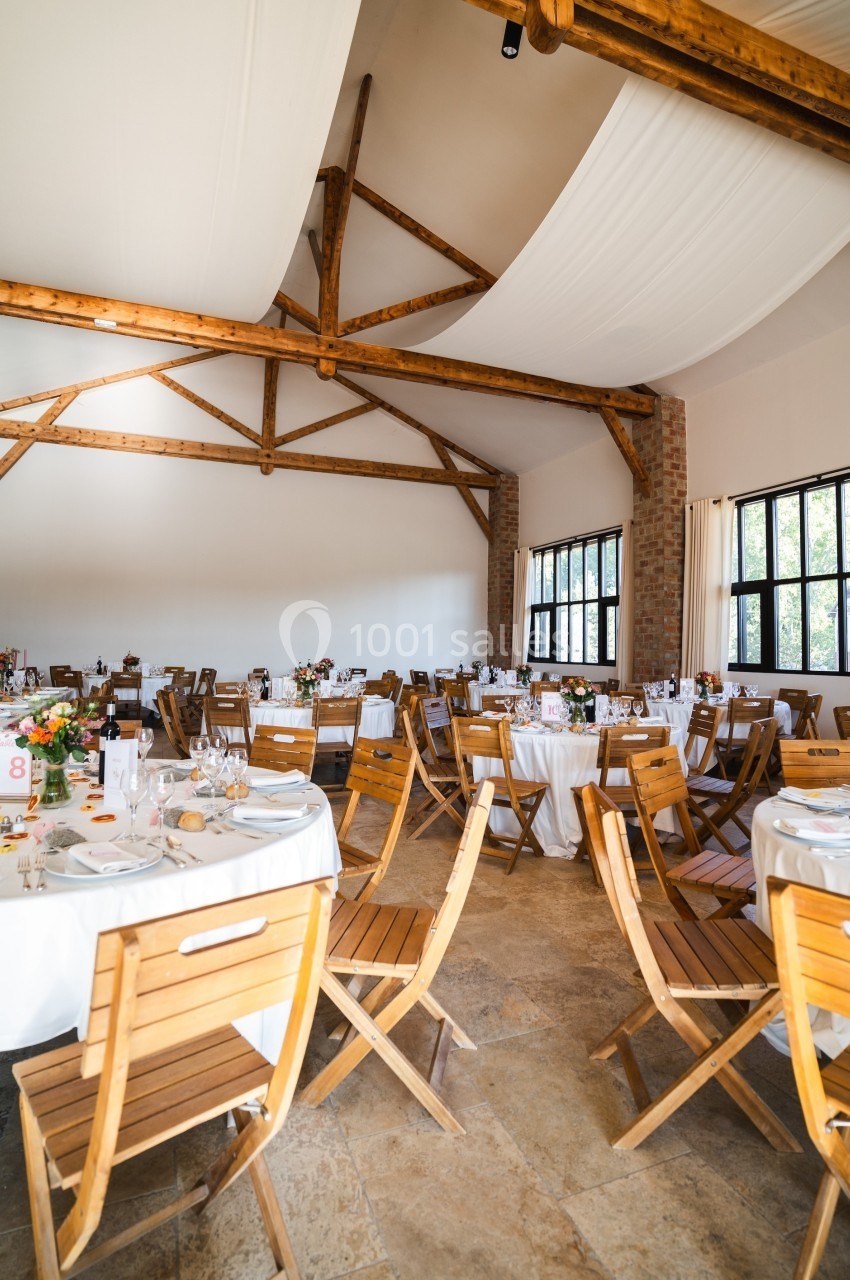 Salle de réception lumineuse avec tables rondes dressées, chaises en bois et poutres apparentes au plafond.