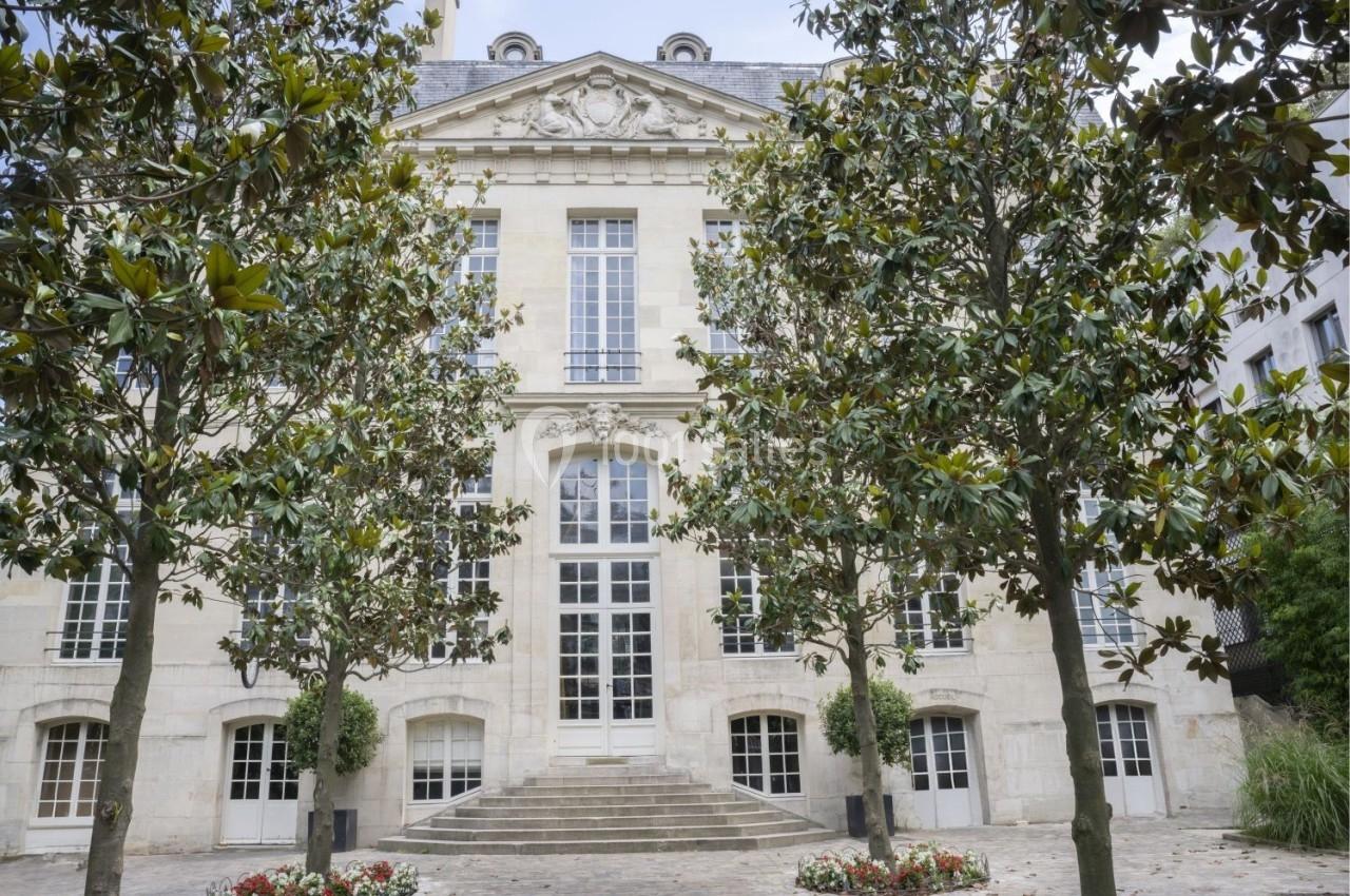 Façade d'un bâtiment en pierre avec des grandes fenêtres, entouré d'arbres et d'un parterre de fleurs.