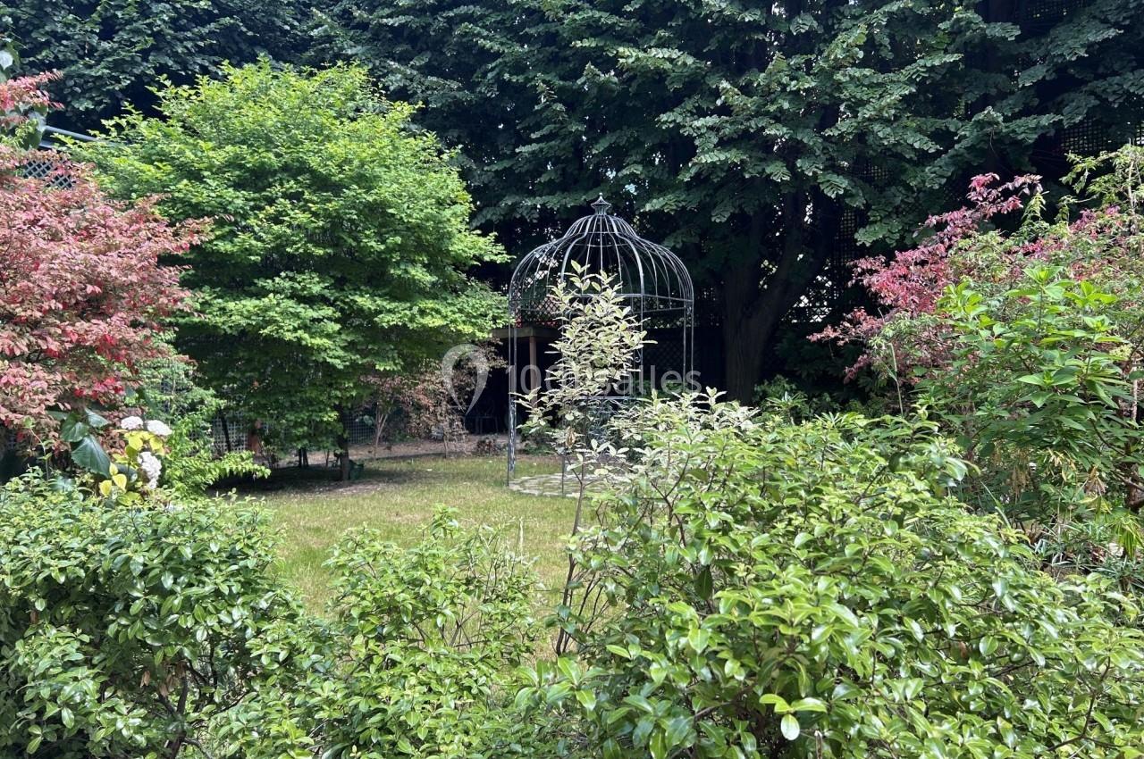 Jardin verdoyant avec une cage en métal décorative au centre, entourée de buissons et d'arbres.