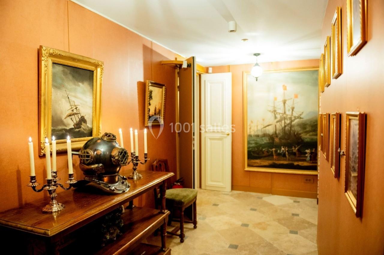 Salle décorée de tableaux maritimes, avec un casque de scaphandrier ancien et des chandeliers sur une table en bois.