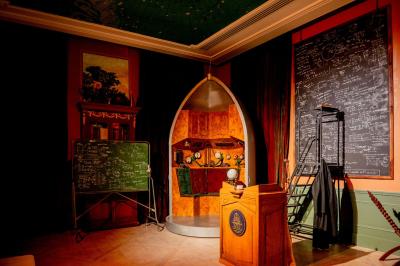 Salle lumineuse avec parquet, tapis décoré, fresques murales, rideaux sombres et plafond orné d'une carte céleste.