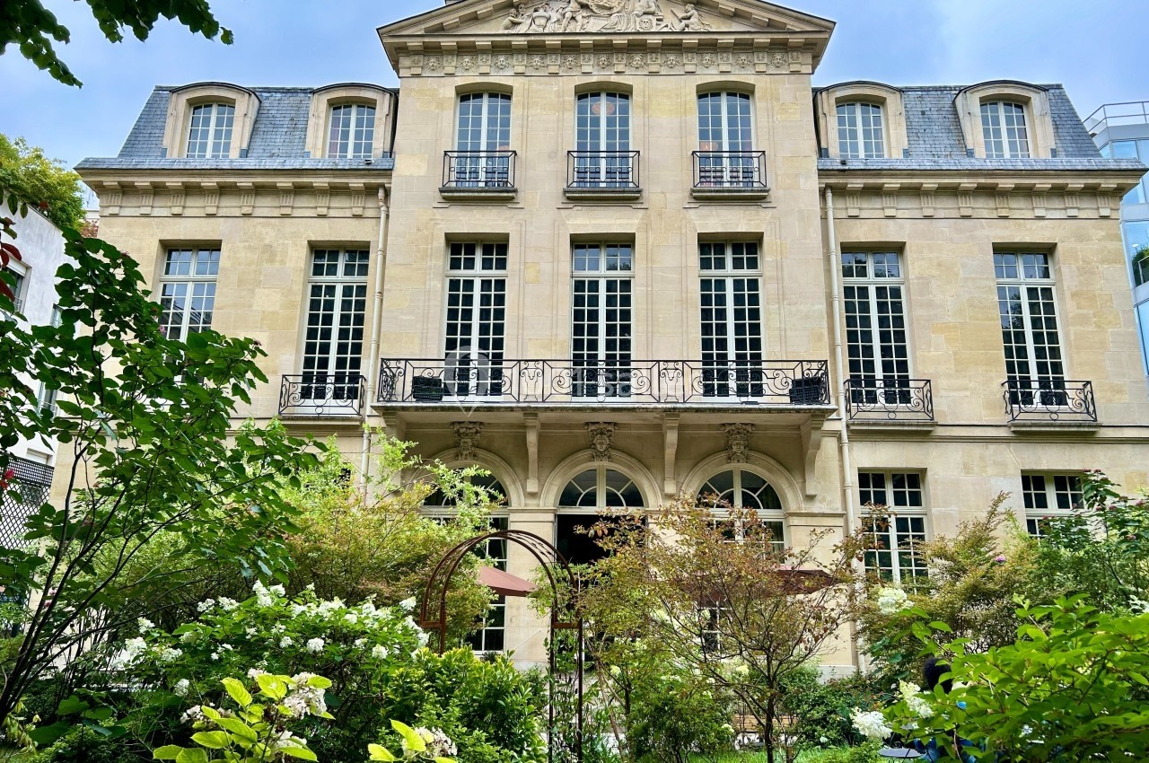 Façade d'un bâtiment ancien en pierre avec grandes fenêtres, vue depuis un jardin verdoyant.
