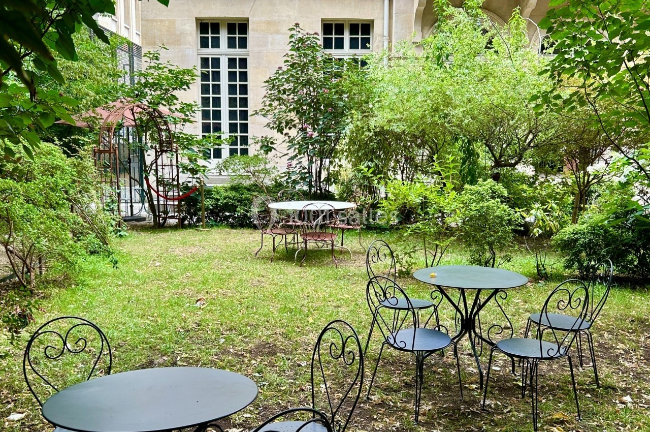 Tables et chaises en fer forgé disposées dans un jardin verdoyant devant une façade de bâtiment ancien.