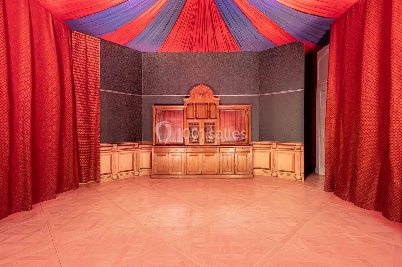 Salle avec parquet clair, murs ornés de rideaux rouges et plafond décoré de drapés rouges et bleus.