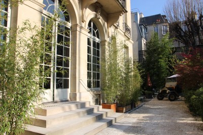 Salle lumineuse avec grandes baies vitrées ouvertes sur un jardin verdoyant, tables et chaises en métal.