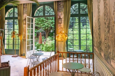 Salle lumineuse avec grandes baies vitrées ouvertes sur un jardin verdoyant, tables et chaises en métal.
