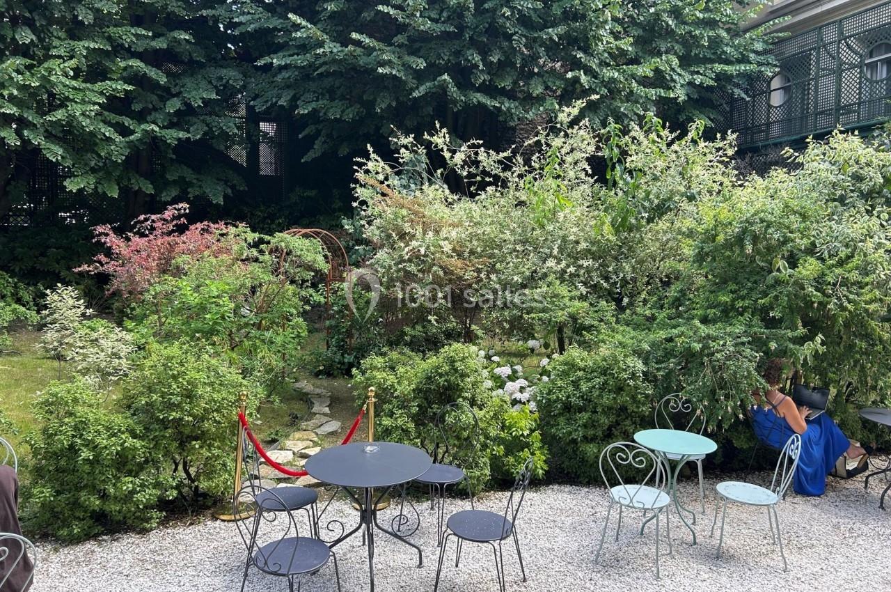 Jardin paisible avec tables et chaises en métal sur un sol gravillonné, entouré de végétation dense et verdoyante.