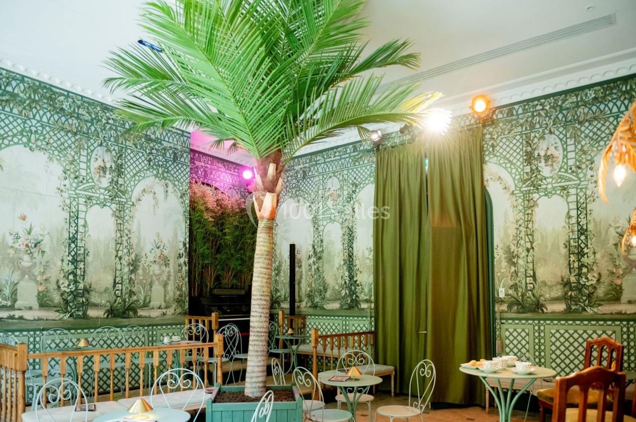 Salle décorée avec des motifs végétaux, un faux palmier central et des tables en fer forgé entourées de chaises.