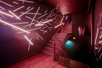 Escalier intérieur avec rampe rouge, éclairé par des néons roses et décoré de motifs lumineux sur les murs sombres.