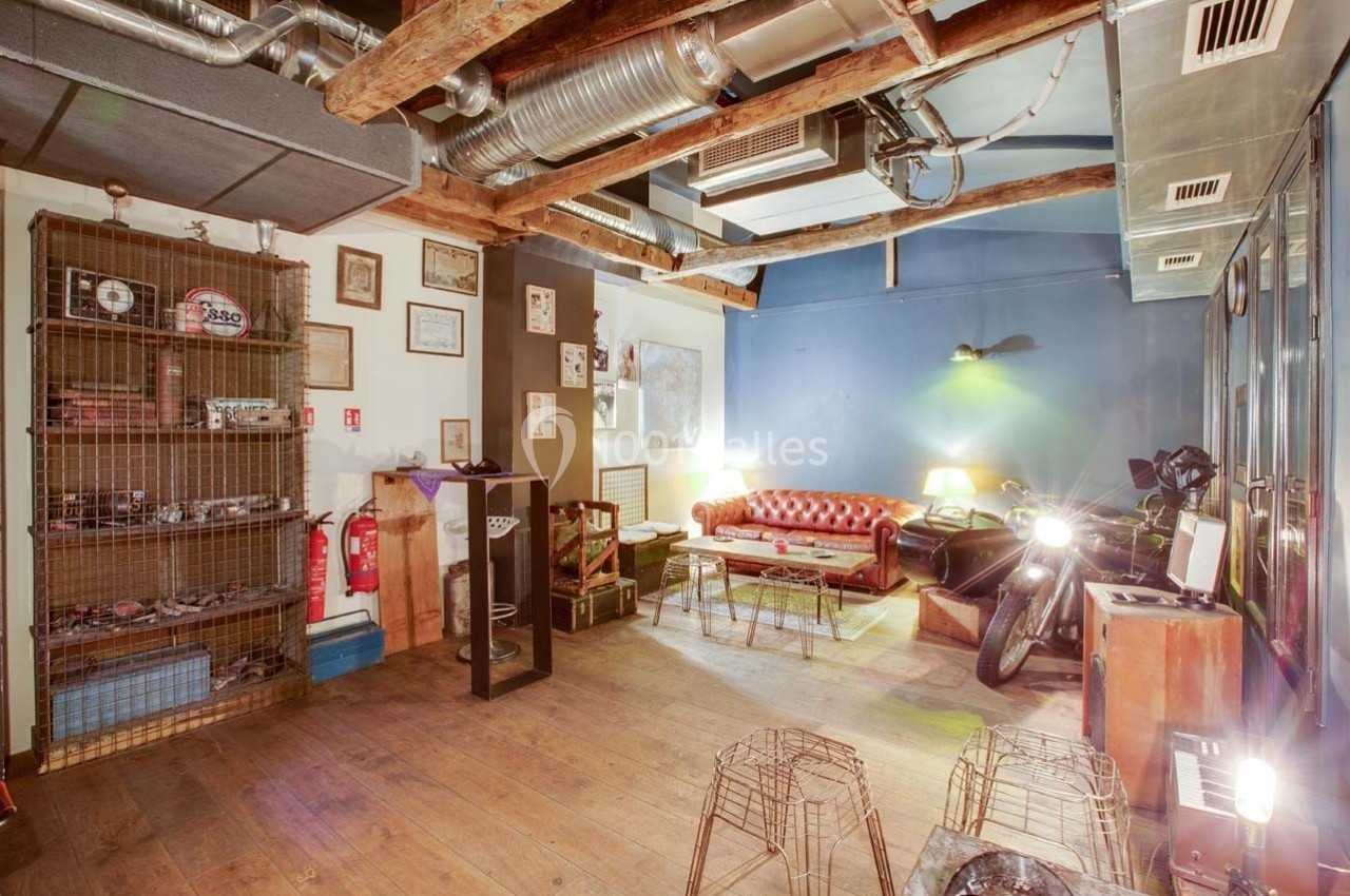 Espace intérieur avec canapé en cuir, moto, étagères métalliques et poutres apparentes dans une ambiance industrielle.