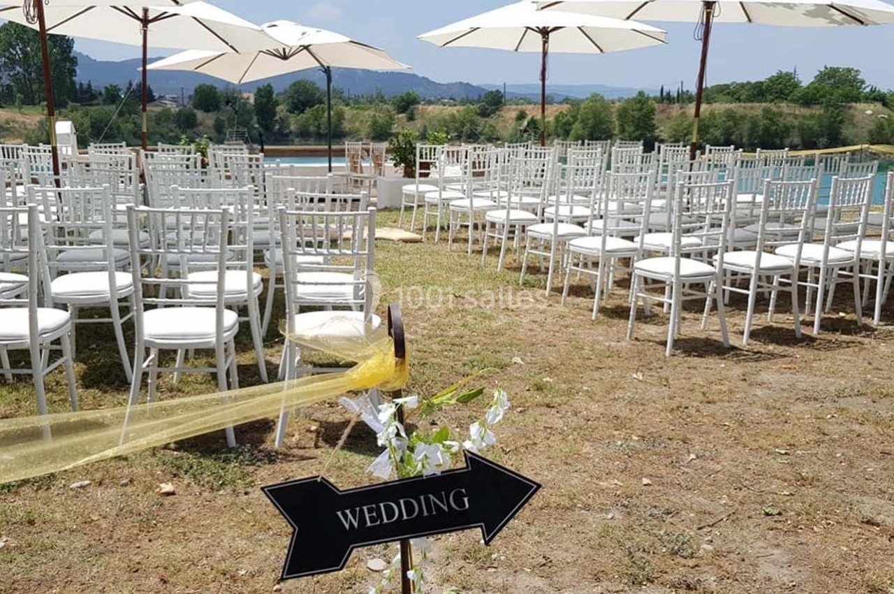 Chaises blanches alignées en extérieur sous des parasols, avec une pancarte indiquant ’Wedding’ et un paysage en arrière…