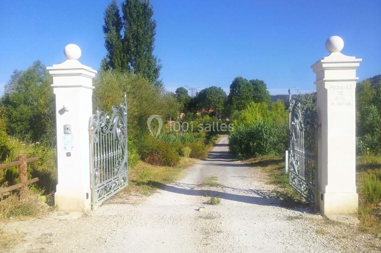 Entrée d'un chemin rural bordé de végétation, encadrée par deux piliers blancs avec des portails en fer forgé.