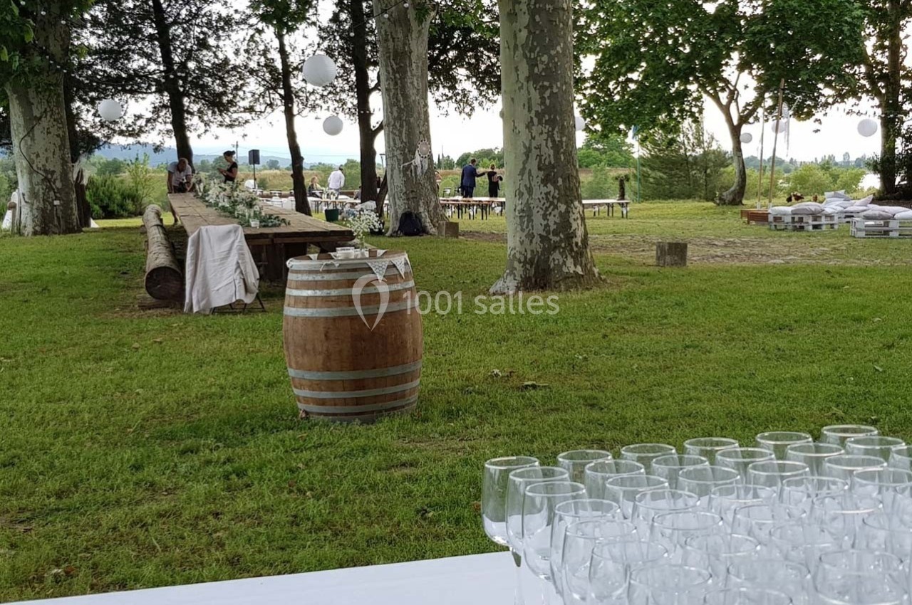 Table dressée avec des verres à pied en premier plan, espace extérieur verdoyant avec tables et décorations en arrière-plan.