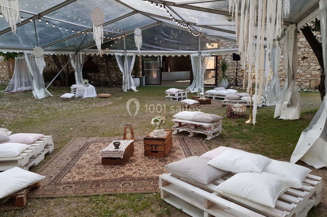 Espace extérieur aménagé sous une tente transparente avec palettes, coussins, tapis et décorations suspendues.