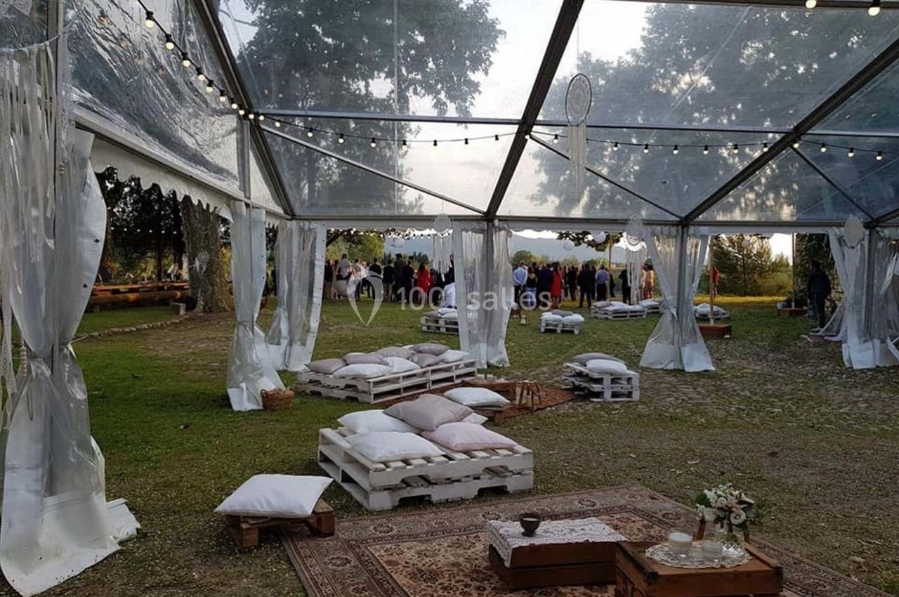 Tente transparente décorée avec des guirlandes lumineuses, coussins et palettes en bois, dans un espace extérieur verdoyant.