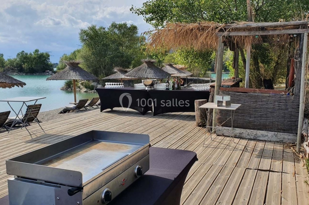 Terrasse en bois au bord d'un lac avec des parasols en paille, une plancha et un espace aménagé pour un buffet.