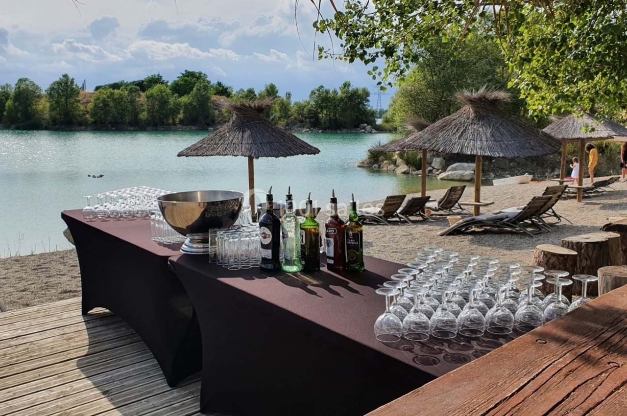Table de bar avec verres, bouteilles et seau à glace, installée en bord de lac avec parasols en paille et transats.
